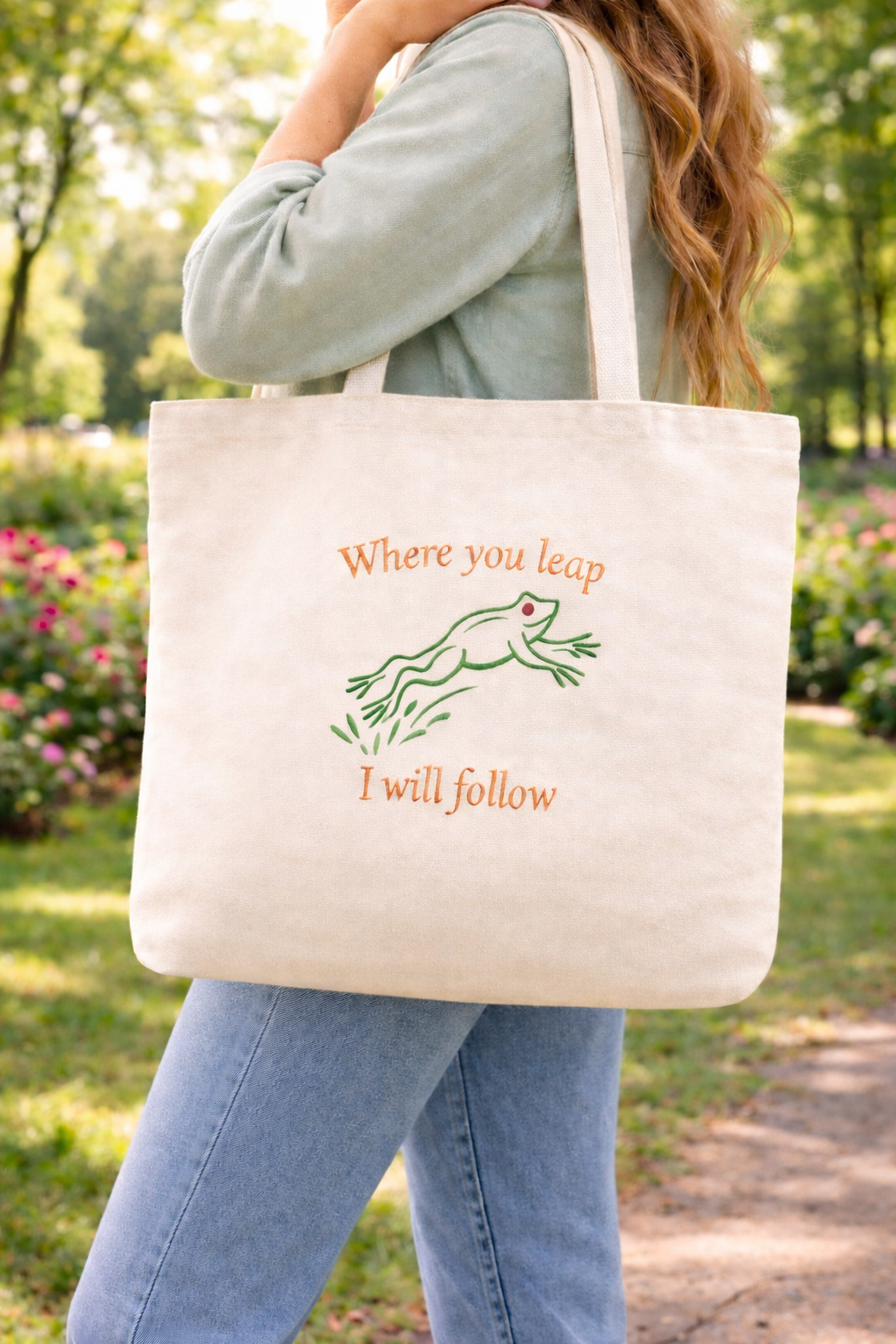 Person holding a canvas tote bag with the embroidered quote "Where You Leap I Will Follow" and a frog design